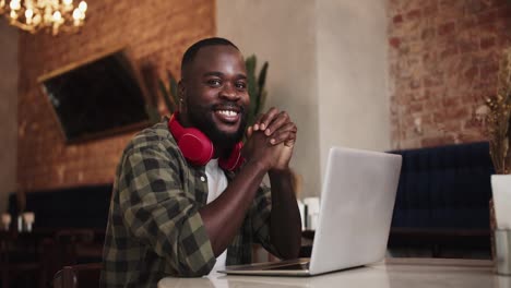 Black-person-folded-his-hands-on-the-table-in-front-of-a-laptop,-posing-and-looking-at-the-camera-while-in-a-cafe