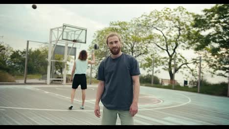 A-red-haired-man-in-a-gray-T-shirt-poses-and-folds-his-arms-on-his-chest-in-front-of-a-team-that-plays-basketball-on-a-basketball-court-in-summer