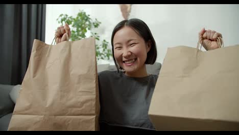 Aufgeregte-Asiatische-Dame,-Die-Online-Einkauft-Und-Zu-Hause-Geschenktüten-über-Ihren-Kopf-Hebt