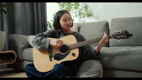 Asian-girl-playing-acoustic-guitar-music-instrument-at-home,-sitting-on-floor