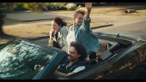A-happy-trio:-a-guy-and-two-girls-are-riding-in-a-gray-convertible.-A-blonde-girl-and-a-brunette-girl-have-fun-and-wave-their-hands-and-look-at-the-camera