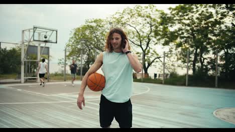 A-curly-haired-red-haired-man-in-a-light-green-T-shirt-and-with-a-basketball-in-his-hands-poses-against-the-background-of-a-team-that-plays-basketball-on-the-basketball-court