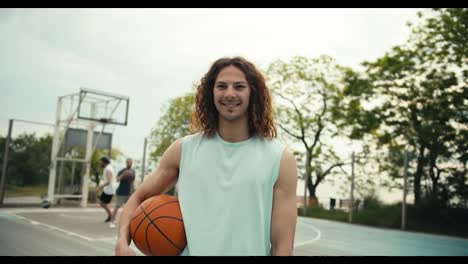 Close-up-shot-of-a-curly-red-haired-man-in-a-light-colored-t-shirt-with-a-basketball-ball-posing-and-looking-at-the-camera-outside-on-a-basketball-court-in-summer