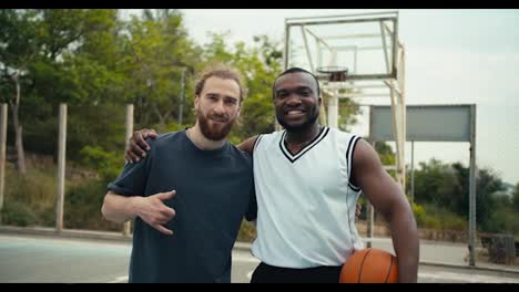 Portrait-of-two-friends,-a-red-haired-man-and-a-black-man-in-a-white-t-shirt,-posing,-looking-at-the-camera-and-smiling-on-the-background-of-a-basketball-court