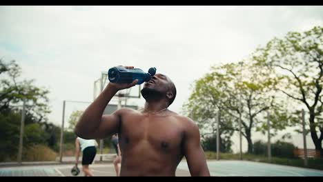A-man-with-Black-skin-and-a-bare-torso-drinks-water-on-a-basketball-court-and-then-licks-her-head-to-cool-off-and-shouts-his-battle-cry-in-front-of-his-friends-playing-basketball