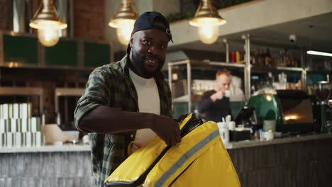 A-food-delivery-man-in-a-plaid-shirt-and-black-cap-poses-and-smiles-at-a-doner-market.-A-man-opens-a-special-thermal-bag-for-delivery