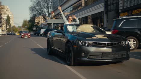 A-glossy-gray-convertible-is-driving-down-the-street,-a-blonde-girl-in-the-back-seat-looks-out-of-the-convertible-and-stretches-her-arms-up