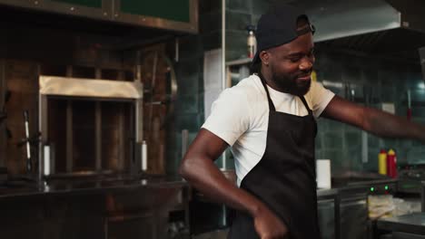 A-Black-person-dances-at-work-in-a-doner-market-against-the-backdrop-of-a-barbecue.-Man-in-black-apron-and-white-t-shirt-having-fun-at-work
