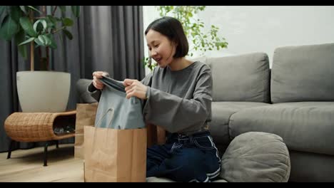 Cute-asian-woman-opening-paper-bag-with-new-clothes-sitting-on-floor-in-living-room-at-home