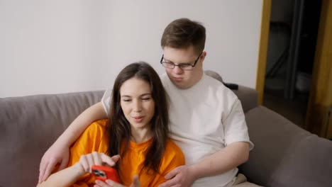 Young-Downs-Syndrome-man-sitting-on-sofa-with-a-girl,-she-is-using-mobile-phone,-scrolling-screen