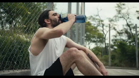 A-middle-aged-man-in-a-white-T-shirt-sits-on-a-basketball-court-near-the-fence,-rests-and-drinks-water-from-a-special-sports-bottle