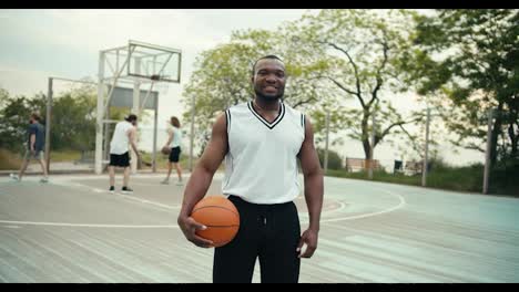 Ein-Schwarzer-Basketballspieler-Mit-Einem-Basketball-In-Der-Hand-Posiert-Und-Blickt-In-Die-Kamera-Vor-Dem-Hintergrund-Seiner-Freunde,-Die-Auf-Der-Straße-Basketball-Spielen