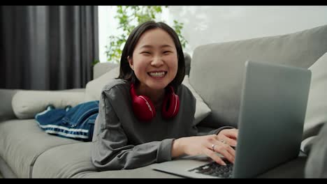 Portrait-of-asian-woman-typing-on-laptop-while-laying-on-sofa,-looking-to-the-camera-and-smiling