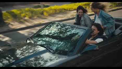 The-happy-trio-ride-in-a-gray-convertible-with-a-breeze.-The-wind-develops-hair,-a-happy-trip-of-young-friends-in-denim-jackets