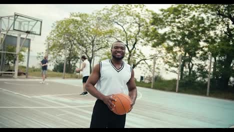 Portrait-of-a-young-Black-man-who-is-posing-looking-at-the-camera-and-smiling-in-front-of-his-friends-who-are-playing-basketball-outside-in-the-summer