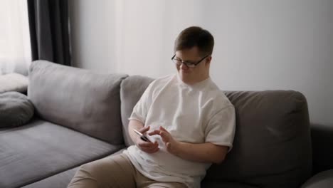Smiling-Downs-Syndrome-guy-sitting-on-sofa-using-mobile-phone,-scrolling-internet