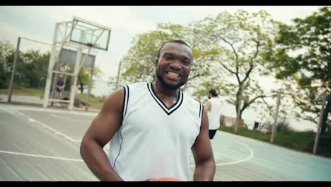 Schwarzer-Mann-Lächelt-Breit,-Posiert-Und-Zeigt-Seine-Gefühle,-Während-Er-Im-Sommer-Mit-Seinen-Freunden-Auf-Dem-Basketballplatz-Basketball-Spielt