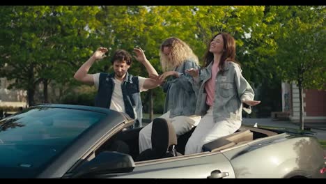 Happy-trio-of-young-friends-in-denim-jackets.-A-guy-and-two-girls-are-dancing-on-the-street-near-their-gray-convertible