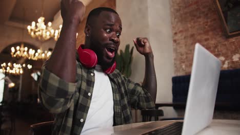 A-black-person-with-a-beard-in-red-headphones-works-on-a-laptop-in-a-cafe,-rejoices-in-success-and-raises-his-hands-up-as-a-sign-of-victory.-Emotions-of-success-at-your-favorite-job