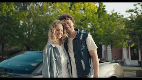 Portrait-of-a-young-brunette-guy-and-a-blonde-girl-who-are-standing-near-their-gray-car-in-denim-jackets-smiling-and-looking-at