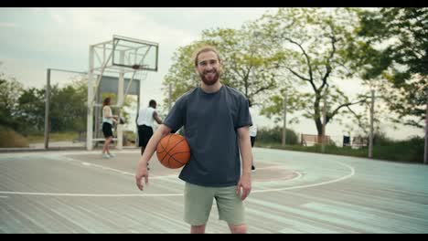 A-red-haired-man-in-a-gray-t-shirt-made-of-an-orange-basketball-poses-against-the-background-of-his-friends-on-the-basketball-court-outside-in-the-summer