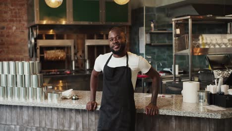 A-Black-person-in-a-black-apron-and-a-white-t-shirt-poses-and-looks-at-the-camera-on-the-background-of-the-doner-market