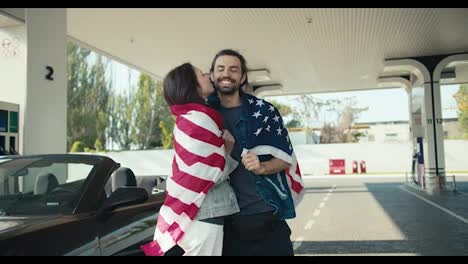 Happy-brunette-girl-kissing-her-boyfriend,-couple-wrapped-in-US-flag-and-standing-at-gas-station-near-their-gray-convertible