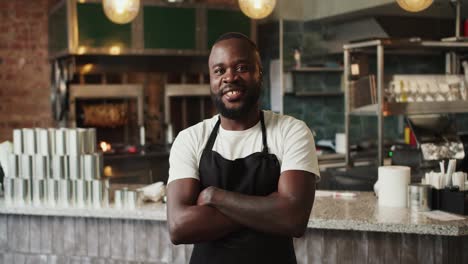 A-Black-person-in-a-black-apron-and-a-white-T-shirt-poses-with-arms-folded-on-his-chest-against-the-background-of-the-doner-market