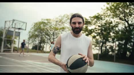 A-middle-aged-man-in-a-white-T-shirt-with-a-beard-poses-against-the-background-of-a-basketball-court-with-a-basketball-in-his-hands