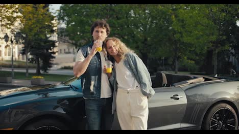 A-guy-and-a-girl-are-standing-near-a-gray-Cabriolet-smiling-and-looking-at-the-camera-and-drinking-yellow-cocktails