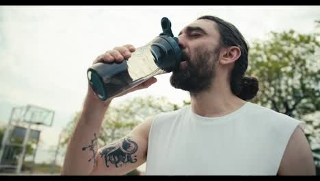 A-middle-aged-man-with-a-tattoo-on-his-arm-and-in-a-white-T-shirt-drinks-water-on-the-basketball-court