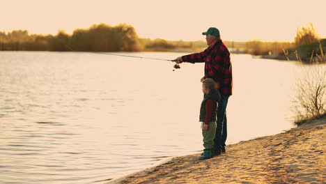 happy-old-man-and-grandson-are-resting-on-river-shore-fishing-from-beautiful-coast-in-sunset-time-active-leisure-and-lifestyle