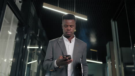 A-black-businessman-walks-down-the-corridor-with-a-mobile-phone-and-looks-at-the-screen