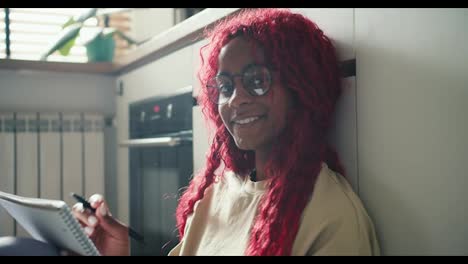 Afroamerikanisches-Mädchen-Mit-Roten-Lockigen-Haaren,-Das-Auf-Dem-Küchenboden-Studiert,-Notizen-Macht,-In-Die-Kamera-Schaut-Und-Lächelt