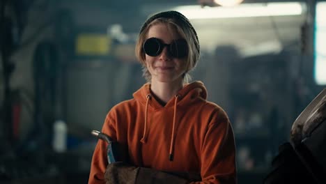 Spark-Your-Passion:-Teenage-Welder-in-Action.-A-blond-teenager-in-goggles-stands-in-a-workshop-in-an-orange-hoodie,-holding-a