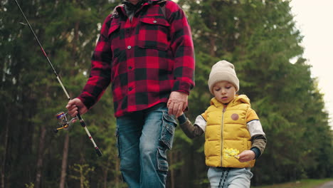cute-little-boy-and-his-granddad-are-walking-together-in-nature-strolling-on-road-across-forest-going-to-fishing