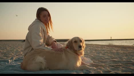 A-blonde-girl-sits-on-a-sunny-beach-with-her-dog-in-a-light-color-in-the-morning