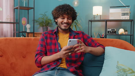 Indian-hindu-young-man-sitting-on-sofa,-using-smartphone-share-messages-on-social-media-application