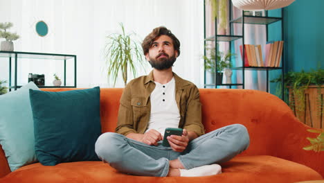 Middle-eastern-young-man-sitting-on-sofa-using-smartphone-share-messages-on-social-media-application