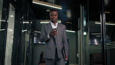 In-the-office-a-black-man-walks-down-the-corridor-with-documents-and-looks-at-the-phone-screen