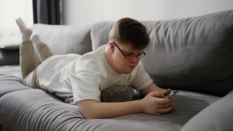 Junger-Mann-Mit-Down-Syndrom-Benutzt-Mobiltelefon,-Während-Er-Sich-Zu-Hause-Auf-Der-Couch-Ausruht