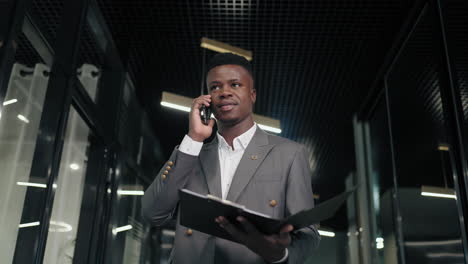Successful-black-business-man-talks-to-smartphone-in-office-interior.-african-american-businessman-walks-down-corridor-and-calls-on-her-mobile-phone.-Portrait-of-friendly-man-walking-down-corridor-.