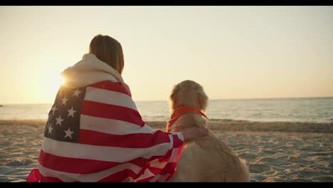 Close-up-shot-A-blonde-girl-sits-on-a-sunny-beach-with-her-dog-wrapped-in-an-American-flag-on-a-sunny-beach-in-the-morning