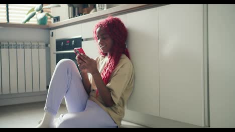 Joyful-African-American-girl-with-red-curly-hair-sitting-on-kitchen-floor-and-srfing-internet-using-mobile