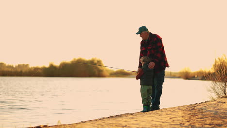old-fisherman-is-teaching-his-grandson-to-catch-fish-in-lake-or-river-in-sunset-time-active-rest-in-weekend