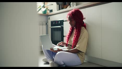 Glückliche,-Erstaunte-Afroamerikanische-Freiberuflerin-Mit-Roten-Lockigen-Haaren,-Die-Mit-Einem-Laptop-Auf-Dem-Küchenboden-Sitzt
