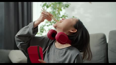 Young-asian-woman-in-headphones-eats-noodles-passionately-with-wood-sticks-at-home