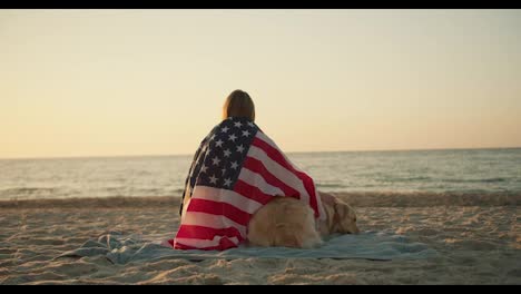 Blondes-Mädchen-Sitzt-Morgens-An-Einem-Sonnigen-Strand-Mit-Ihrem-Hund,-Der-In-Eine-Amerikanische-Flagge-Gehüllt-Ist