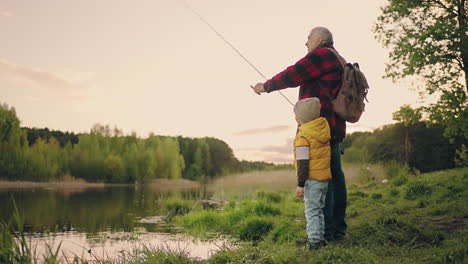 old-man-and-little-boy-are-fishing-on-shore-of-picturesque-lake-in-forest-grandfather-and-grandson