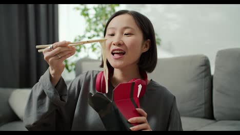 Young-asian-woman-in-headphones-eats-noodles-with-wood-sticks-at-home